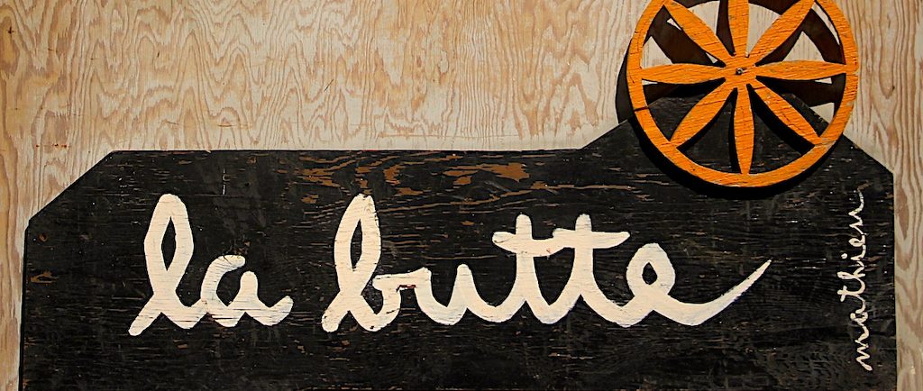 Color photo of the Butte sign on which we can read La Butte on a dark background surmounted by an orange cart wheel.