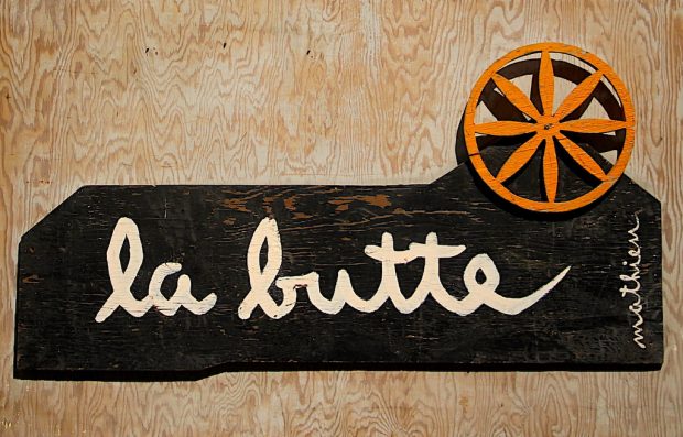 Color photo of the Butte sign on which we can read La Butte on a dark background surmounted by an orange cart wheel.