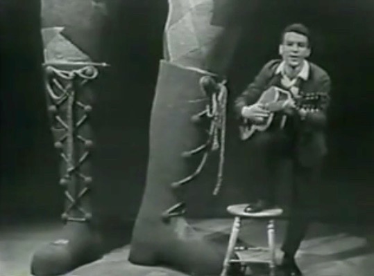 Black and white photo of Claude Gauthier accompanying himself on the guitar. A pair of lumberjack boots appears in the foreground.