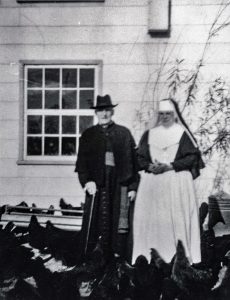 Black and white photo of a man wearing a black tunic and hat, accompanied by a nun wearing a veil, apron, and cross. They are surrounded by hens.