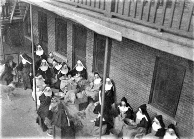 Black and white photo of a group of women wearing nuns’ habits and aprons. They are peeling broad-beans, backs to the convent.