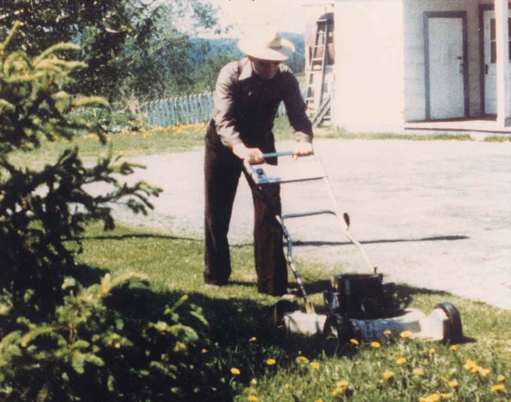 Colour photo of a man wearing a hat and mowing the lawn.