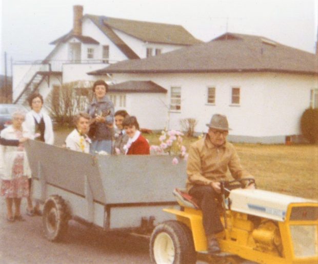 Colour photo of a man sitting on a small yellow tractor pulling a grey trailer where a few women are sitting. One of the women is holding a guitar. They are in front of the farmhouse.