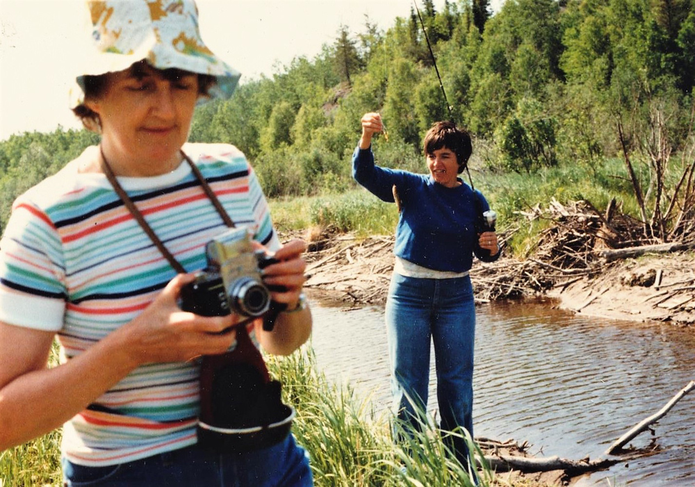 Photo en couleur de deux femmes en plein air près d’une rivière. L’une pêche tandis que l’autre tient un appareil photo entre ses mains.