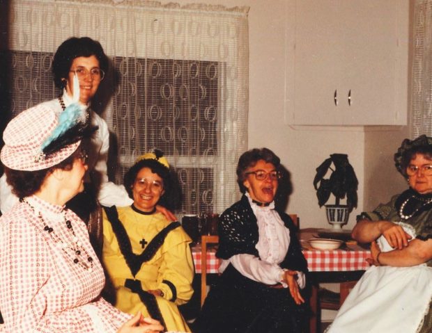 Colour photo of five women wearing colourful dresses and hats in a kitchen.