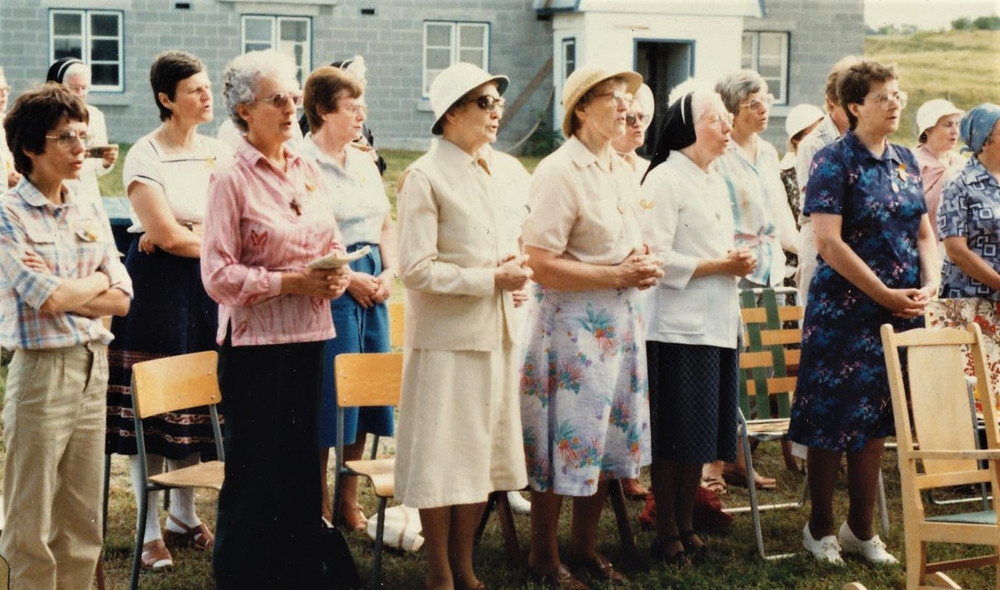Photo of women in front of the barn. Several are wearing hats. Their hands are joined.