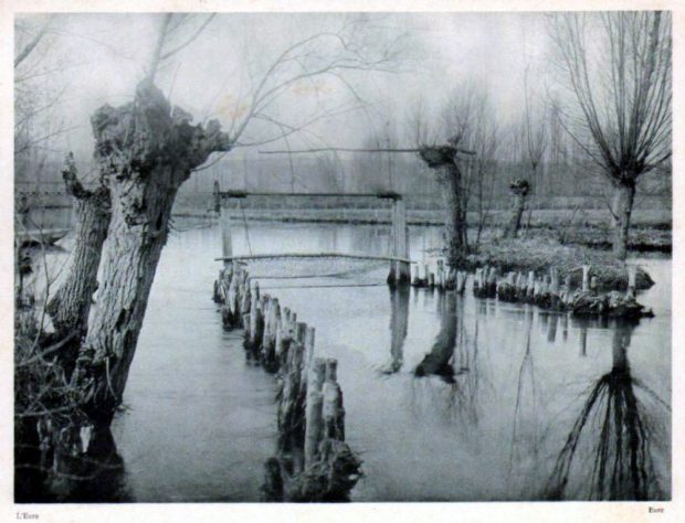 In a river bordered by trees that have lost their leaves, two parallel rows of short posts form a passageway that is closed off at the end by a net. Black and white photograph.