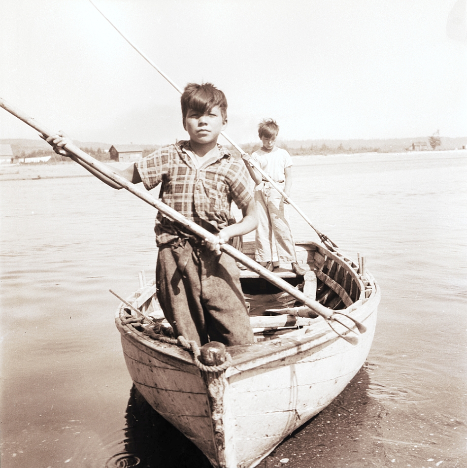 Two Aboriginal boys stand in a wooden rowboat near the shore, facing the photographer. Each one is holding a nigogue, a type of harpoon, above the water as if preparing to spear a fish. Black and white photograph.
