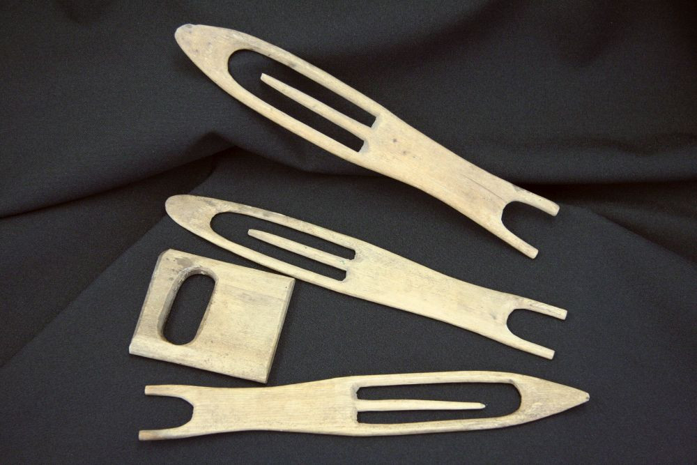 Four hand-crafted tools made of thin pieces of wood, lying on black fabric. They consist of three needles with a rounded point for knitting fishing nets and a small square object that serves as a mesh gauge.