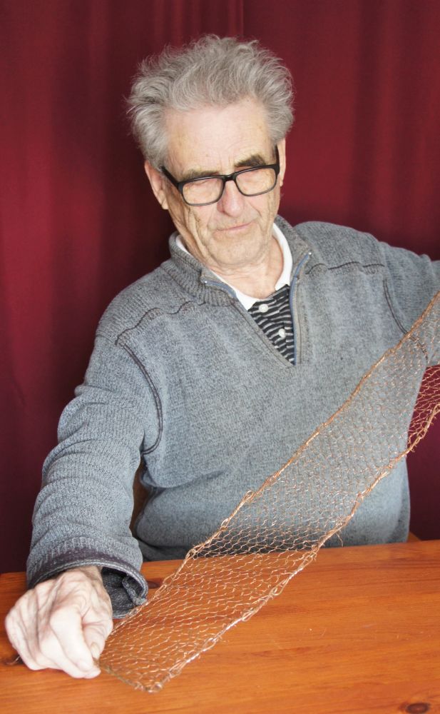 A man in his eighties sitting at a table holds a narrow strip of copper netting between his hands.