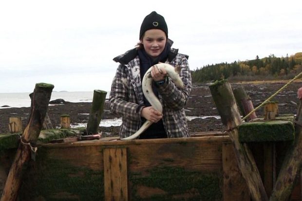 A teenage girl wearing a tuque and a coat stands in a wooden box used to hold eels captive in a weir. She is holding an eel in her hands. The river and a cove can be seen in the background.