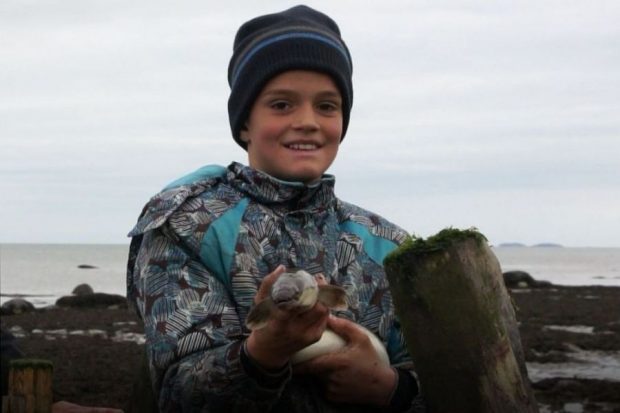 Head and shoulder view of a young boy dressed in winter clothing, proudly holding an eel in his hands. He is showing the eel’s head. The river can be seen in the background.