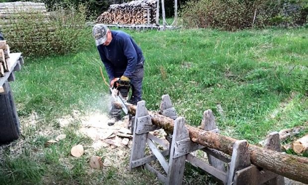 A man uses a chain saw to shape into a point the end of a post lying on a home-made sawbuck.