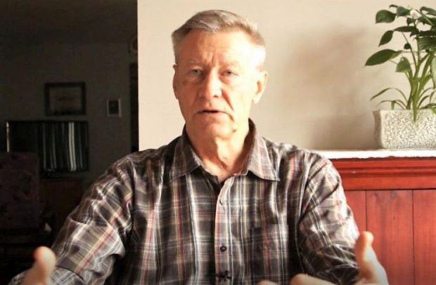 Close-up view of a man in a checkered shirt sitting behind a desk or a table and looking sideways.