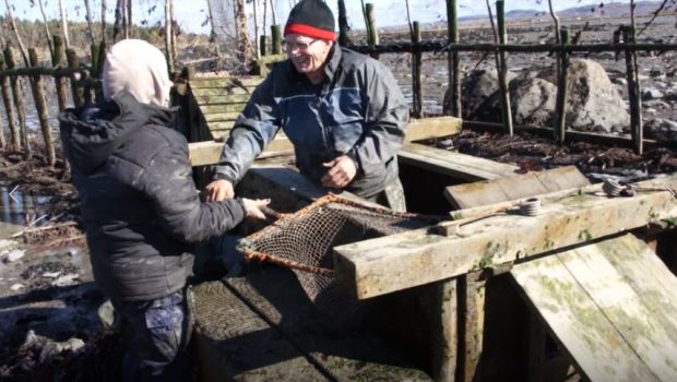 A man standing in a wooden box used to hold eels captive in the weir takes a net mounted on a square frame with a wooden handle from a woman who is handing it to him.