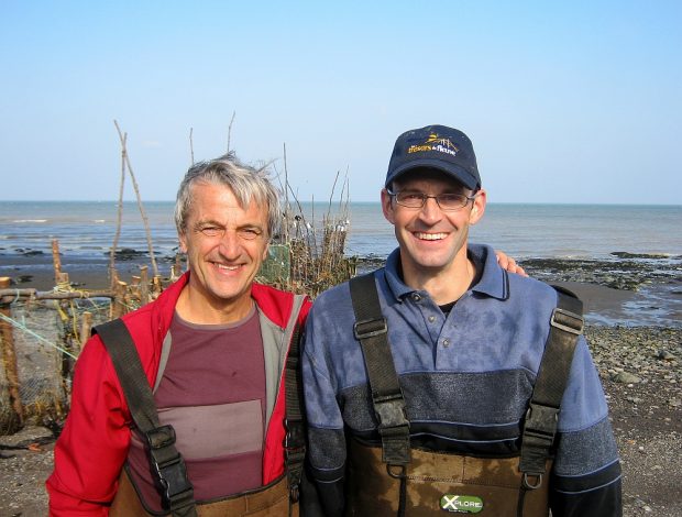 Head and shoulder view of two men returning from eel fishing, with their arms around each other’s shoulders. An eel weir and the river can be seen in the background.