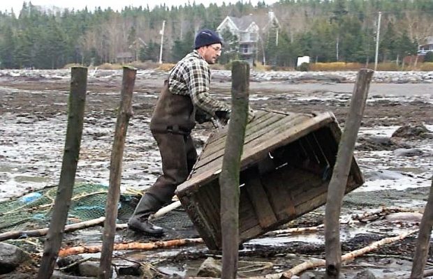 A man moves a large wooden funnel on the shore of the river by rolling it on the ground.