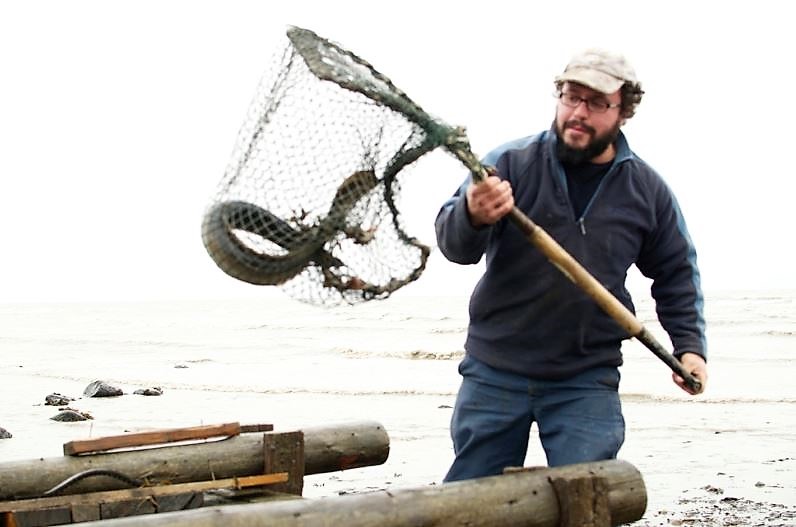 An eel fisher holds a large net attached to a rigid frame with a handle, containing an eel. The top of a large wooden collecting box can be seen at the bottom of the photograph.