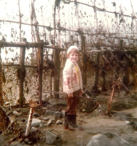 In this photograph from the 1970s, a young boy wearing rubber boots, a jacket and a wool hat smiles as he poses in front of an eel weir with vertically installed fishing nets. He has his feet in the water.