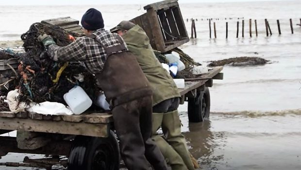 Two men in fishing clothes put a net on a trailer, whose wheels are sitting in the incoming tide.