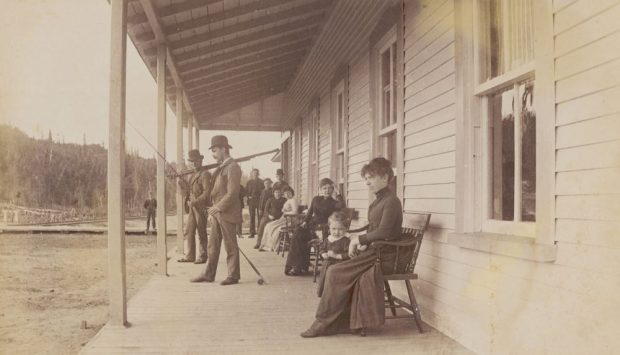 Sepia-toned photo of a group of people on the porch of the private club known as Laurentide House. The women and children are seated, while the men are standing with their fly- fishing rods. All are elegantly attired.