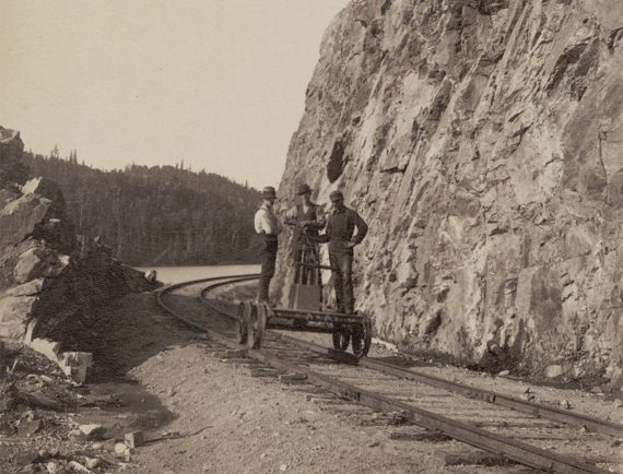 Photo en sépia présentant trois hommes en chariot ferroviaire et un talus rocheux sur le lac Édouard.  Inscription en bas de la photo Rock cut lake Edward.