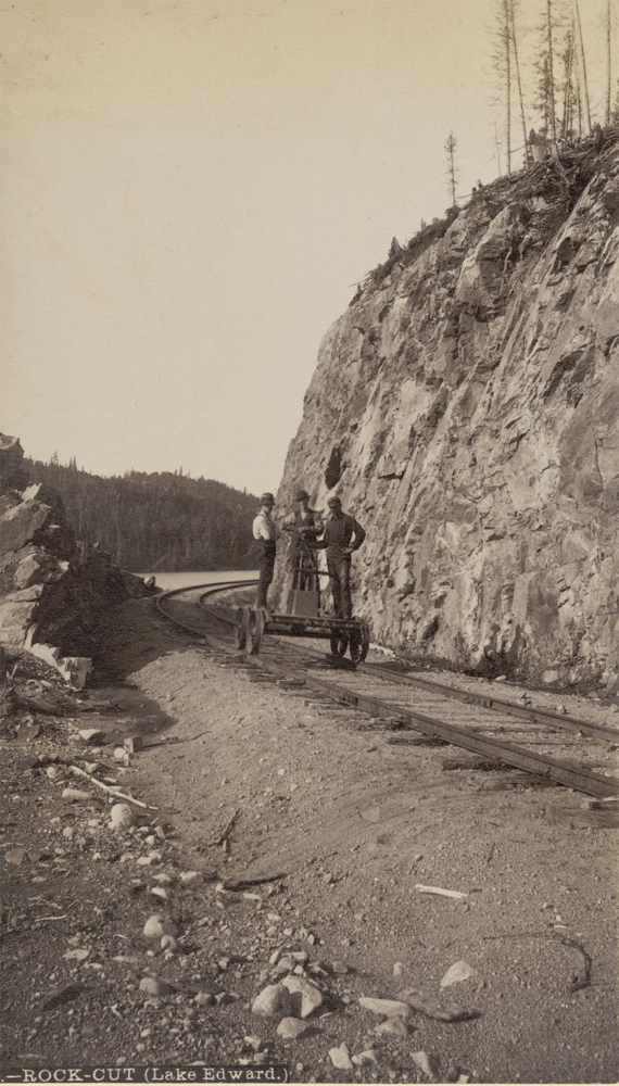 Sepia-toned photo of three men in a railway carriage and the rocky banks of lac Édouard. Inscription at bottom of photo reads: Rock cut Lake Edward.