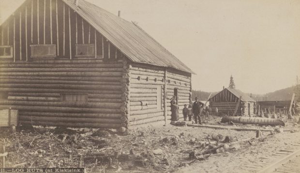 Sepia-toned photo of a log cabin; in the entrance stands a family looking off in the direction of the railroad that passes close by. In the background lie other homes.