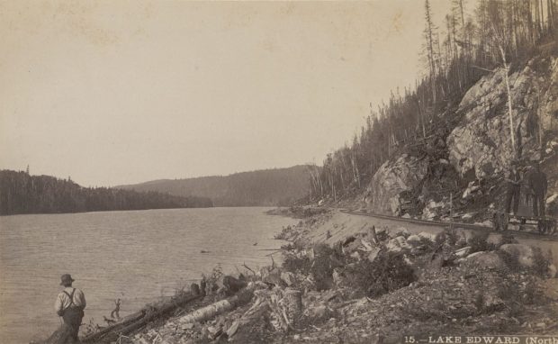 Sepia-toned photo shows how close the railroad is to lac Édouard. Workers on a railway carriage; in the foreground someone fishes on the banks of lac Édouard. Inscription at bottom of the photo reads: Lake Edward (North).