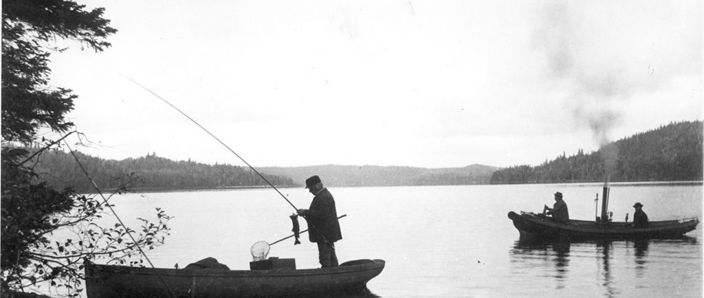 Black and white photo of fishermen in a steamboat on lac Édouard. In the foreground, a boat on the river’s edge; in it stands a man unhooking a fish from his rod.