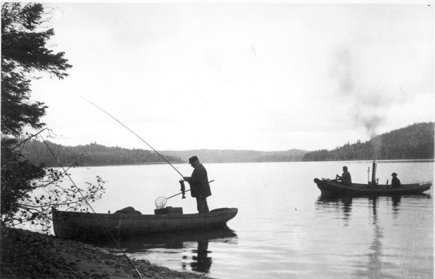 Black and white photo of fishermen in a steamboat on lac Édouard. In the foreground, a boat on the river’s edge; in it stands a man unhooking a fish from his rod.