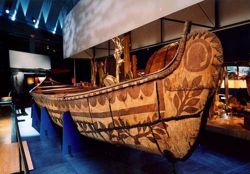 Colour photo, a 36-foot canoe made from birch bark and embellished with engravings. The canoe rests on a base and is displayed in the Musée de la civilisation de Québec.