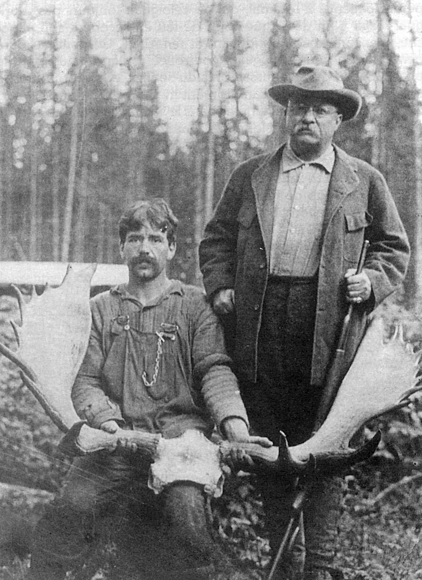 Black and white photo, two men proudly pose with impressive moose antlers.