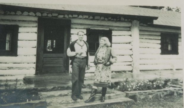 Photo en noir et blanc,  Un homme, Fowler McCormick et une femme Anne Stillman sont debout devant un chalet en bois rond.