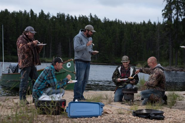Photo en couleur de 5 hommes dégustant un bon repas en plein air sur les berges d'un plan d'eau.
