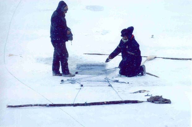 Color photo, two people are on a frozen lake. A hole is drilled in the ice and a fishing net is nearby. Two wooden poles will hold the net in place.