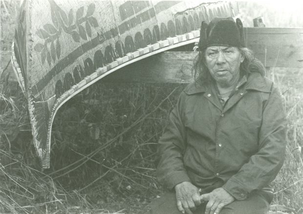 Black and white photo, César Newashish sitting next to a white birch bark canoe. Floral and ancestral motifs adorn the boat’s hull.