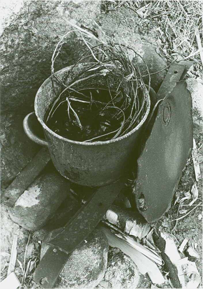 Photo en noir et blanc, préparatif pour l'assouplissement de racines d'épinette.  Sur des pierres est déposé un chaudron contenant de l'eau et les racines.