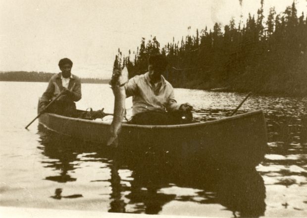 Sepia-toned photo, two men in a boat. The man seated near the front of the boat is holding a pike.