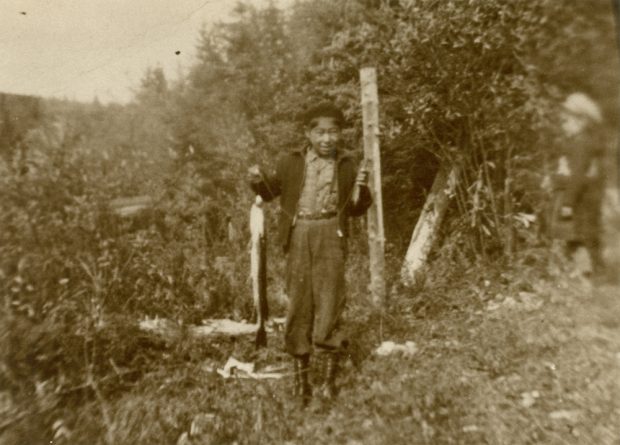 Photo sépia, sur le bord d'un boisé un enfant atikamekw tient dans une main un brochet.