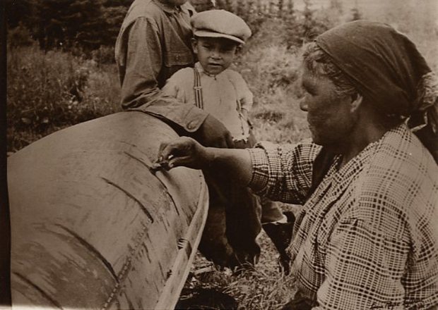 Photo sépia, en avant plan, une femme atikamekw applique de la gomme de sapin sur un canot en écorce de bouleau sous les yeux attentifs d'un jeune garçon