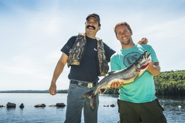 Photo en couleur, deux hommes debout tenant une grosse truite grise, en arrière-plan un lac.