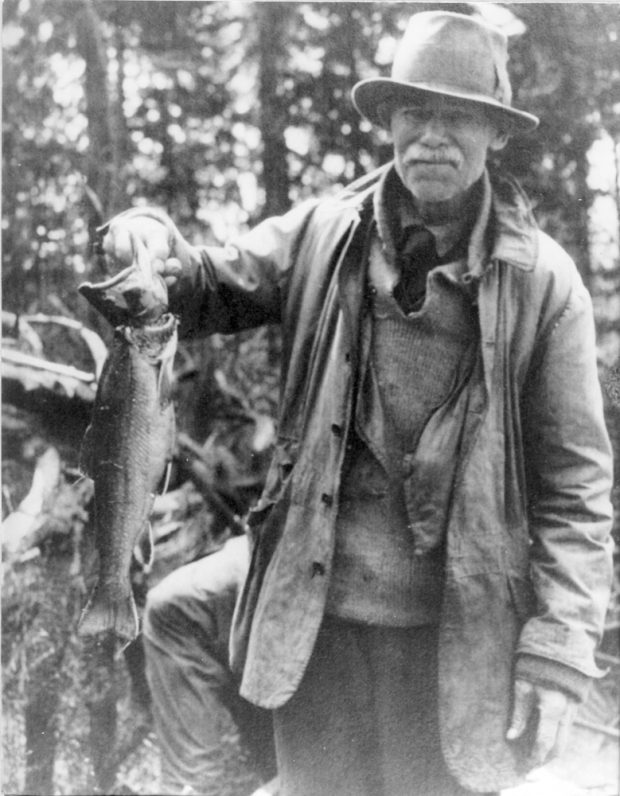 Photo en noir et blanc, un homme debout tenant dans sa main une belle truite mouchetée.