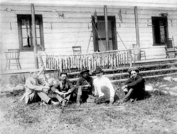 Black and white photo, a group of fishermen in front of the private club on lac Wayagamak, in the background several fish hang from a wooden pole.