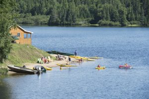 Colour photo, a cottage stands just a few metres from a lake, a group of adults and children enjoy water sports: swimming, kayaking, paddle-boating, and pedal-boating. A few Adirondack chairs are installed on the beach, where vacationers sit and enjoy the moment.