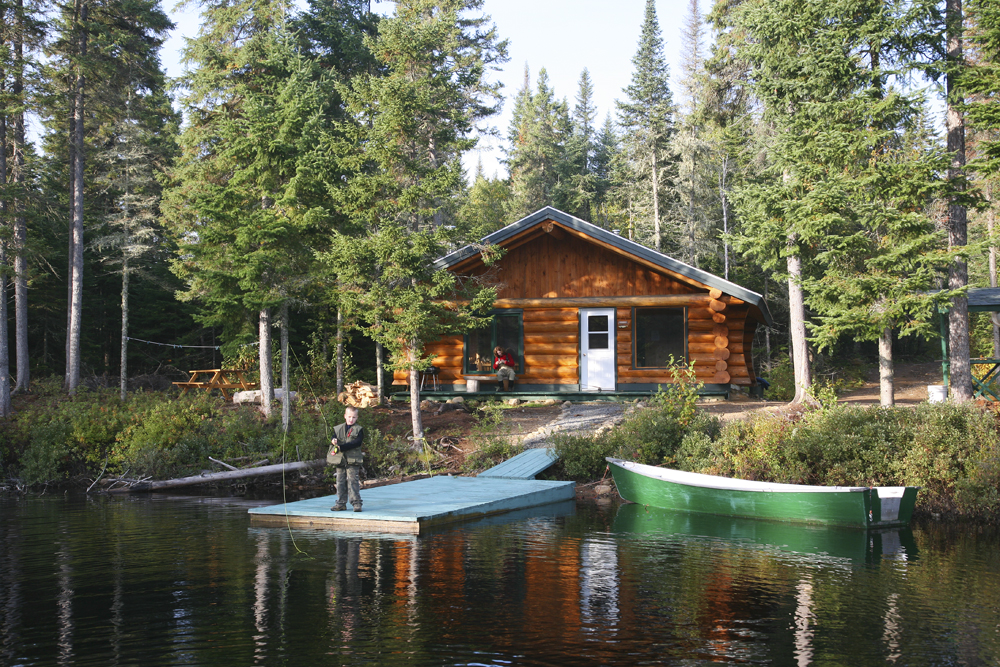Colour photo, a log cabin with a lake just a few metres away. A child standing on the edge of a dock practices casting with a fly-fishing rod. A woman looks on from the balcony. Serene atmosphere, all is calm.