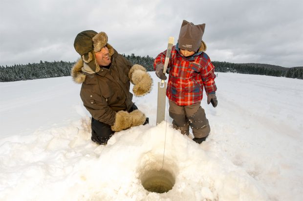Photo en couleur, un père heureux d’initier et son jeune fils à la pêche blanche.  Un trou est percé dans la glace et une brimballe est installée, ils sont prêt pour la pêche sur glace.