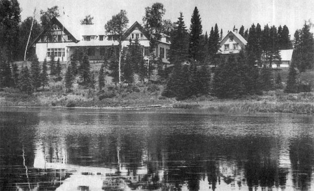 Black and white photo of a stylish house on the edge of a river. A smaller house on the right is partly hidden behind soaring spruce trees.