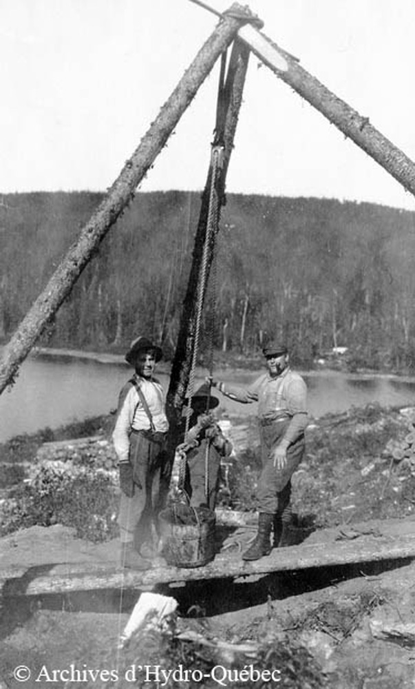 Black and white photo, three workers. Three logs bound together support a pulley that helps retrieve clean drinking water.