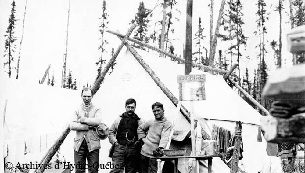 Black and white photo, three workers in front of a prospector camp. Canvas tent with wooden logs as its structure. In front of the tent, a wooden sign with the word Office.